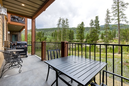 A covered outdoor patio with a black table and chairs, a barbecue grill, and a scenic view of trees and hills in the background.