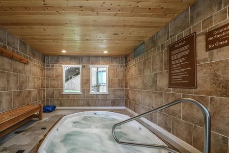 Indoor hot tub with bubbling water, tiled walls, a wooden bench on the left, and rule signs on the right. Natural light enters through two small windows. A handrail leads into the tub.