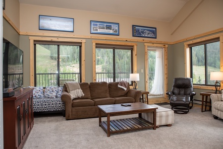 Spacious living room with brown sofa, armchair, wooden coffee table, and scenic artwork. Large windows and glass door provide views of greenery outside. A TV is mounted in the corner.