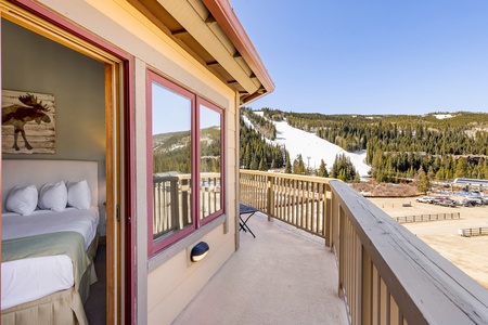 A bedroom with an open sliding door leads to a balcony that overlooks a ski resort, including slopes and forested hills. The room has a bed with white pillows and linens, and a moose painting on the wall.