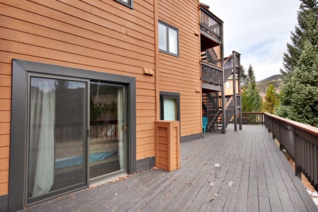 A wooden balcony with railings attached to a multi-story building with sliding glass doors, leading to the interior. Trees and a mountainous background are visible.