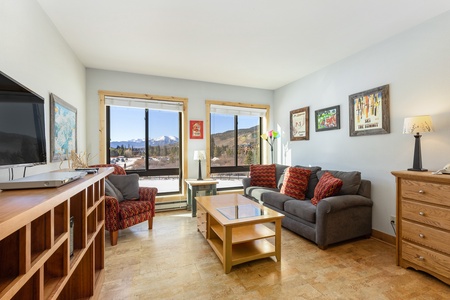 A living room with large windows displaying a mountain view. The room has a couch, armchair, coffee table, TV, and wooden furniture. Framed art hangs on the walls. Sunny and well-lit.