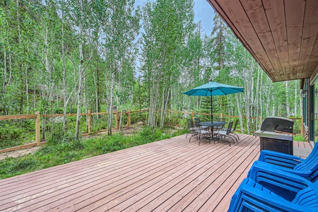 A wooden deck with a table, chairs, blue umbrella, and blue chairs overlooks a fenced forest area with tall trees. A grill is positioned near the sliding door.