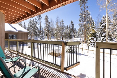 A view from a snow-covered deck featuring two chairs. The scene includes trees, a cabin, and a clear, sunny sky.