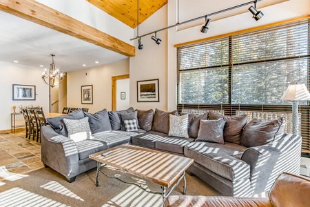 A spacious living room with a large grey sectional sofa, a coffee table, and a window with wooden blinds. In the background, there's a dining area with a long table and a chandelier.