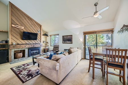 A cozy living room with a beige couch, a mounted TV above a fireplace, a dining table with chairs, and large windows showing a view of trees.