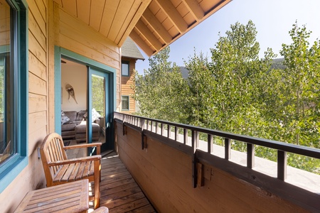 A wooden balcony with two chairs overlooks green trees. A partially open door leads inside to a living room with a couch.