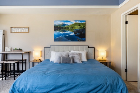 A bedroom with a blue comforter on a bed, a painting of a lake and mountains above the headboard, lamps on nightstands, and a small desk with a Keystone, Colorado sign on the wall.