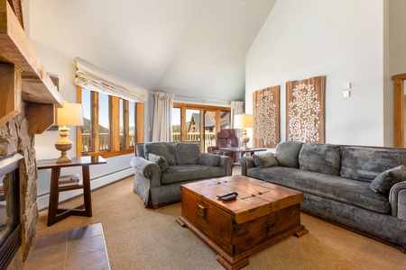 A living room with high ceilings, two sofas, a large wooden coffee table, a stone fireplace, and a window offering a view of the outdoors. The room is decorated with lamps and wall art.