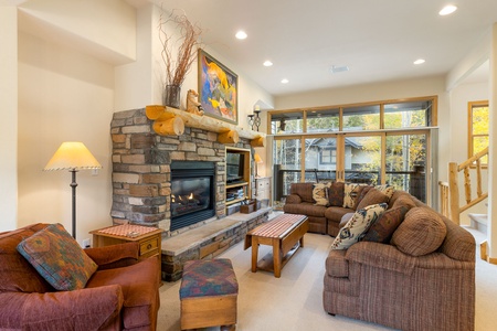 A cozy living room with stone fireplace, two brown sofas, an armchair, wooden coffee tables, and large windows revealing an outdoor view.