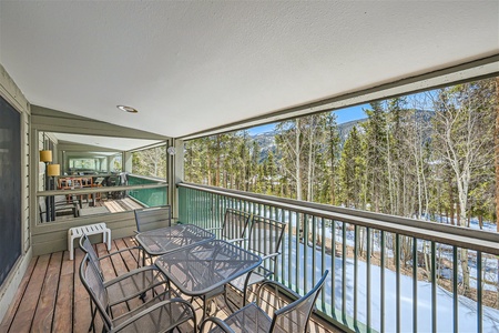 A covered balcony with a table and chairs overlooks a snow-covered landscape and forest area. Another part of the balcony features additional seating and a railing.