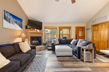 A cozy living room with a fireplace, flat-screen TV, sectional sofa, and a wall adorned with a snowy mountain photo. Light wooden doors and a large window are present in the background.