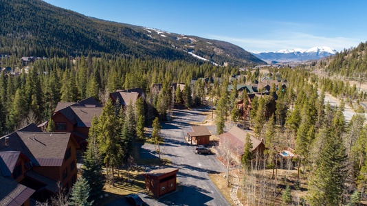 Aerial view of a residential area with wooden houses surrounded by forested mountains under a clear blue sky. Snow-capped peaks are visible in the distance.