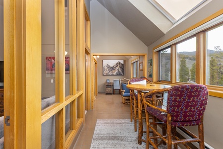 A cozy room with large windows overlooks a forested landscape. The space includes a small wooden dining table with four rustic chairs and a carpet on the tiled floor. A mountain painting is on the wall.