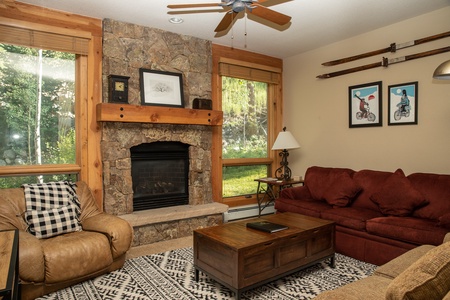 A cozy living room with a stone fireplace, brown sofa, leather recliner, and a wooden coffee table. The room is decorated with framed art and vintage skis on the wall. Large windows offer natural light.