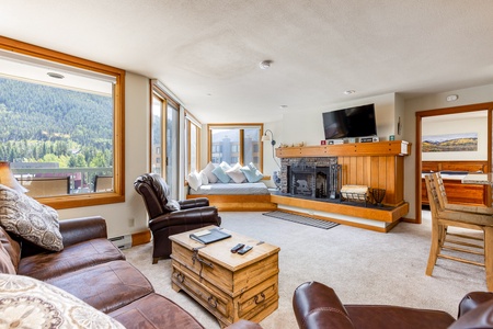 A living room with a leather couch, armchairs, a wooden coffee table, a fireplace, and a television. Large windows provide a view of the mountains and let in natural light.