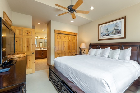 A bedroom with a large wooden bed, white linens, and an overhead ceiling fan. Visible in the background is a bathroom with a double sink and large mirror. The room has wooden furniture and doors.