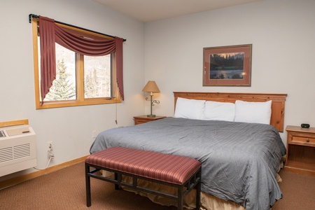 A hotel room features a king-sized bed with a gray duvet, two nightstands with lamps, a bench at the foot of the bed, a window with red drapes, and a landscape painting on the wall.