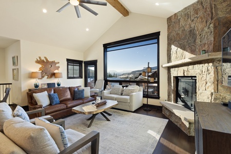 A modern living room featuring a large window with a scenic mountain view, a brown leather sofa, a white armchair, a wooden coffee table, and a stone fireplace.