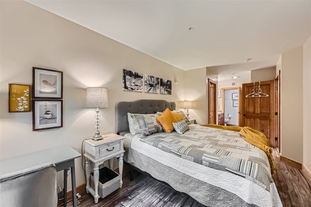 A small bedroom with a large bed, gray tufted headboard, and patterned bedding. The room has a desk, a nightstand with a lamp, framed art on the walls, and a wooden door leading to another room.