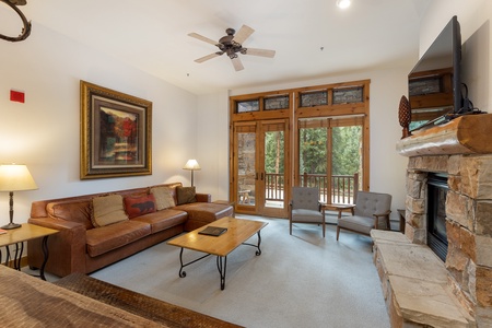 A cozy living room with a leather sofa, two armchairs, a wooden coffee table, a mounted TV, and a stone fireplace. Large windows and a glass door provide a view of a forested area outside.