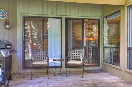 A wooden deck with two metal chairs and a round table in front of sliding glass doors, adjacent to a gas grill. The deck is part of a house with large windows.