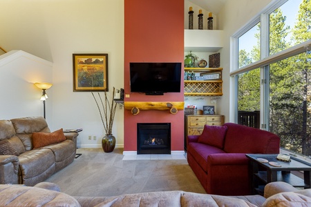 A cozy living room with a red accent wall, a fireplace, a TV above it, brown and red sofas, and large windows displaying a forest view. Shelving and decorations are present on the right side.