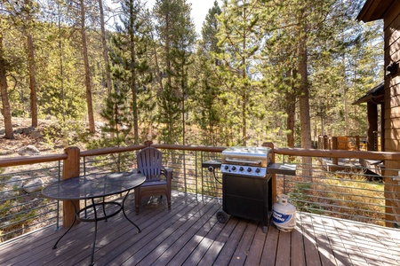 Wooden deck with a round table, plastic chairs, and a gas grill. Surrounded by tall trees and natural forest scenery.