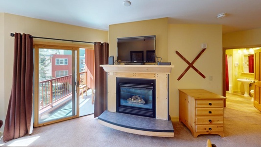 A cozy room with a gas fireplace, mounted TV, wooden chest of drawers, and sliding glass door leading to a balcony. Brown curtains flank the door, and a pair of crossed ski poles adorns the wall.