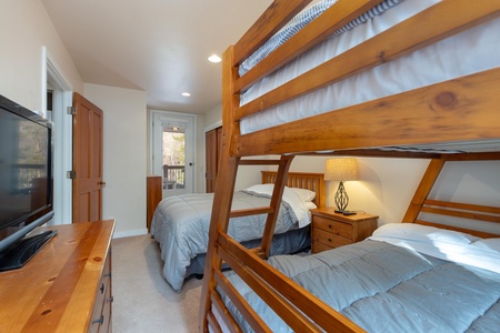 Bedroom with a bunk bed and a single bed, wooden furniture, a table lamp, and a TV. Natural light coming from the door with a window. Light beige walls and carpeting.