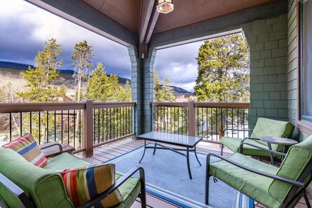 A wooden patio with green cushioned chairs, a coffee table, and a rug overlooks a scenic view of trees, mountains, and a cloudy sky.