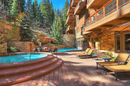 Outdoor patio with hot tub, swimming pool, lounge chairs and umbrellas, bordered by tall trees and a multi-story stone building on a sunny day.