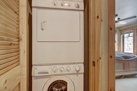 A stacked washer and dryer unit is positioned in a compact laundry area with wooden framing, adjacent to a partially visible bedroom with a bed, window, and ceiling fan.