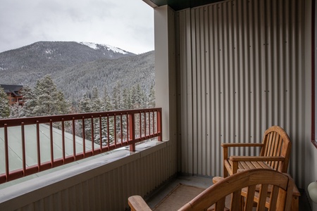 A wooden bench and chair sit on an apartment balcony with a view of snow-covered trees and mountains in the background.