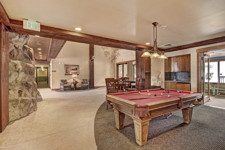 A recreational room featuring a pool table with red felt, a stone accent wall, seating area, wooden furniture, and large windows allowing natural light. An exit sign is visible above a door.