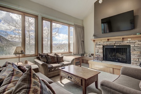 A cozy living room with large windows, beige sofas, a wooden coffee table, and a stone fireplace with a TV above it. Natural light brightens the space, revealing a scenic outdoor view.