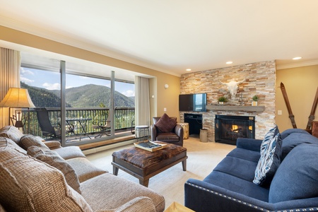 A cozy living room with a stone fireplace, TV, and large windows overlooking a mountain view. It features a sofa, armchair, coffee table, and sliding door leading to a balcony.