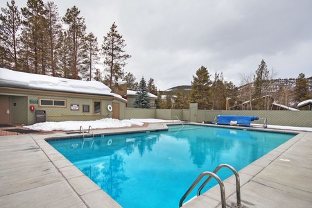 Outdoor swimming pool with metal ladders on either side, surrounded by a concrete deck. Snow-covered trees and a small building are in the background under a cloudy sky.