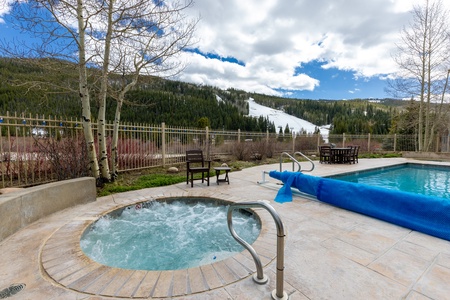 Outdoor hot tub and swimming pool with lounge chairs on a patio, set against a backdrop of snowy mountain slopes and a clear blue sky.