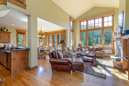 A spacious living room with large windows, wooden flooring, and a mix of leather and fabric furniture. The room features a mountain view, a stone fireplace, and an adjoining dining area and kitchen.