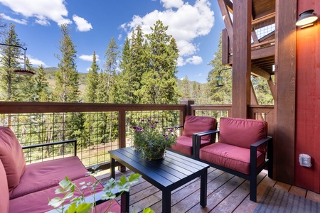 Outdoor patio with red cushioned chairs and a black table, surrounded by wooden fencing. Background of trees and blue sky with clouds.