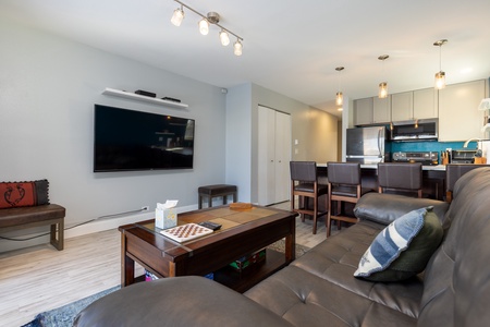 Modern living room with a wall-mounted TV, a leather sofa, a coffee table, and an adjacent kitchen area featuring a countertop with bar stools and stainless-steel appliances.