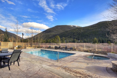 A rectangular outdoor pool with adjoining hot tub, surrounded by a patio with chairs and tables, set against a backdrop of forested mountains under a partly cloudy sky.