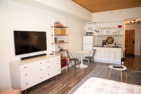 A modern studio apartment with a flat-screen TV on a dresser, open shelving with kitchen items, a small dining table with two chairs, and a kitchenette equipped with appliances.