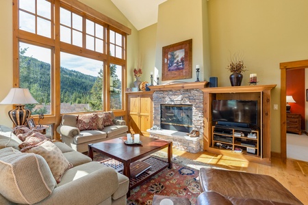 A cozy living room with large windows, mountain views, stone fireplace, comfortable seating, wooden furniture, a rug, and a TV.