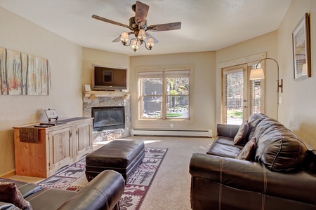 A cozy living room features a leather sofa and armchairs, a stone fireplace, a wall-mounted TV, and large windows with a view outside. A wooden cabinet and an ottoman complete the setting.