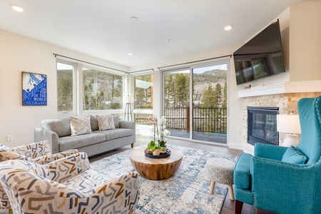 A living room with large windows, mountain views, a gray sofa, a patterned armchair, a teal chair, a round wooden coffee table, and a wall-mounted TV near a fireplace with a painting above it.