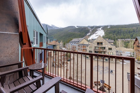 View from a balcony showing a snowy mountain village with several multi-story buildings, a ski slope in the distance, and a wooden chair in the foreground.