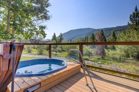 A wooden deck features a round hot tub with metal stairs, overlooking a lush green landscape and distant mountains under a clear sky.