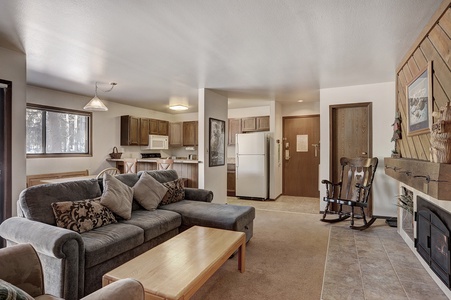 A cozy living area features a gray sofa with cushions, a wooden coffee table, a rocking chair by the fireplace, and an adjacent open kitchen with wooden cabinets and standard appliances.